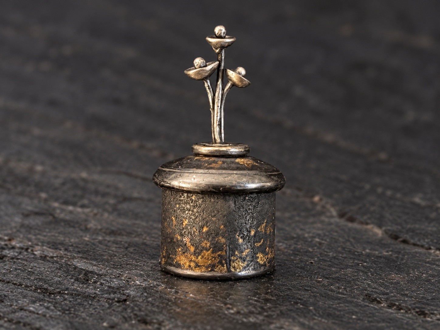 Textured silver box with gold accents and a delicate sculptural stem, photographed against a dark background